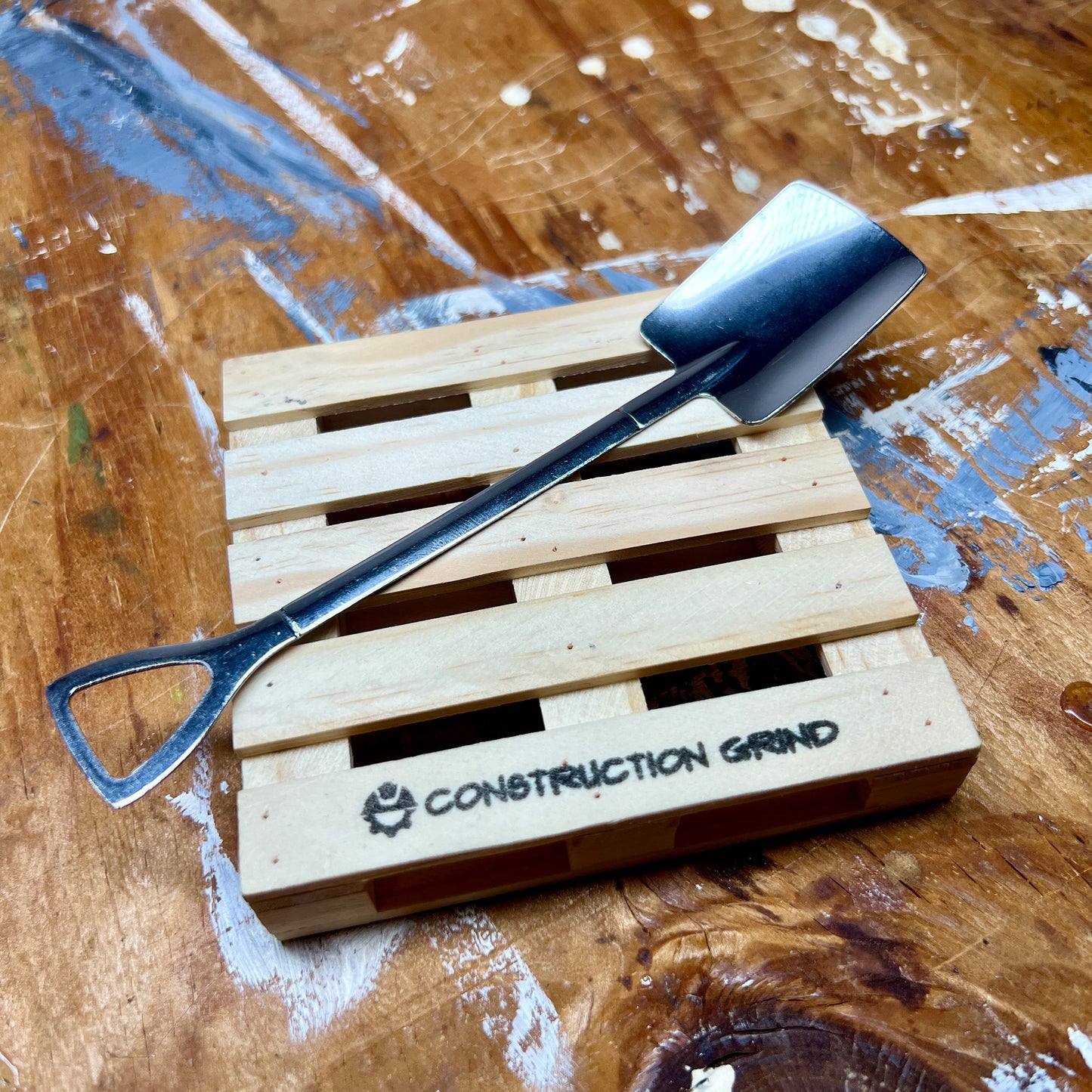  A closeup of Construction Grind's 4" square pallet coaster with a hand stamped Construction Grind logo, and stainless-steel shovel spoon laying on the pallet.